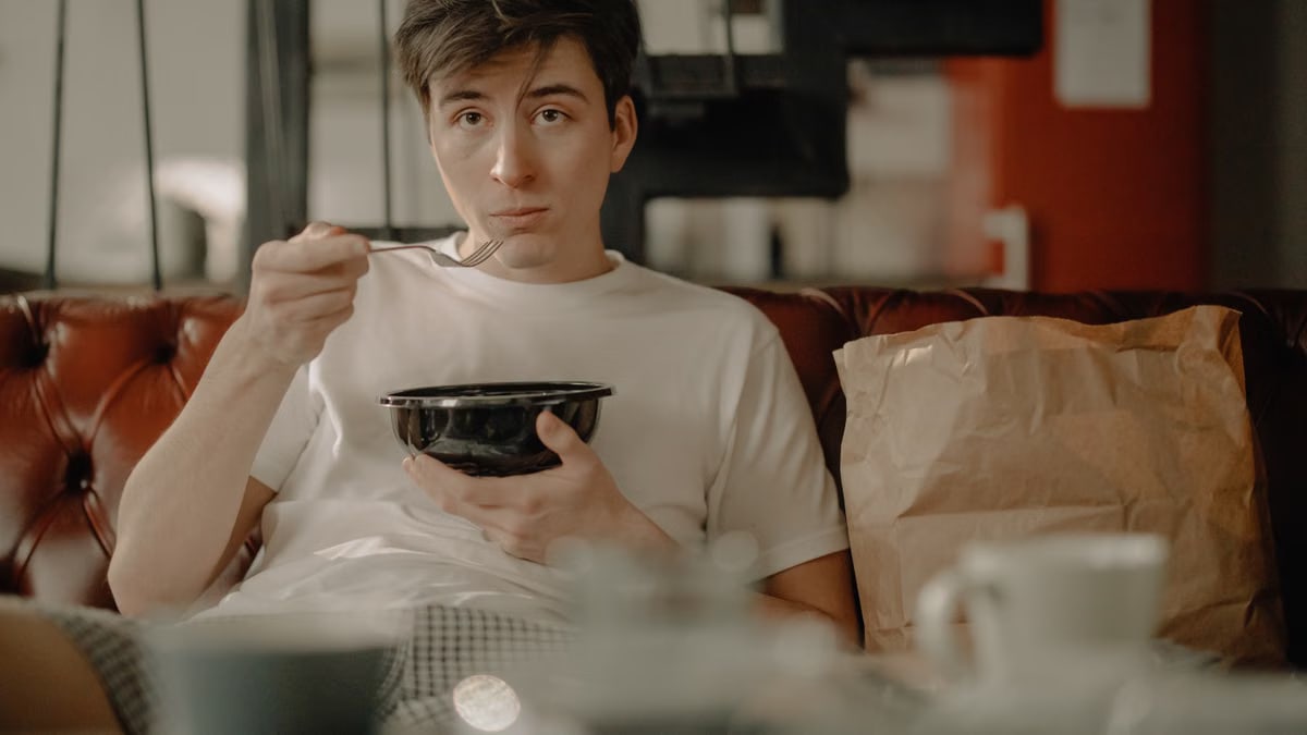 A young man sitting on a couch in an apartment, enjoying his meal with a paper bag nearby.